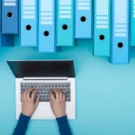 Photo of a person typing on a laptop next to archive boxes.