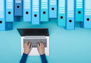Photo of a person typing on a laptop next to archive boxes.